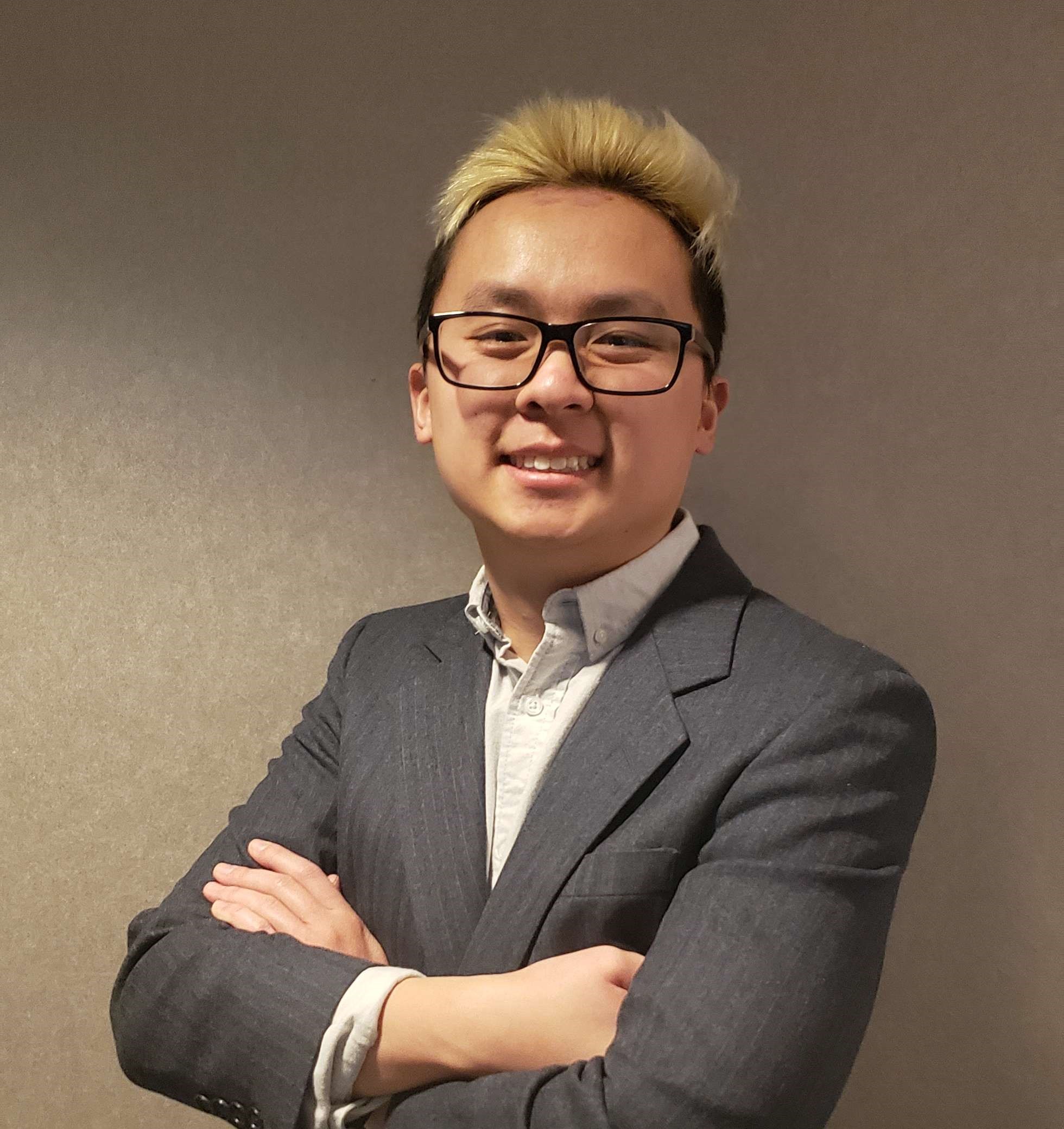 A portrait of me against a wall wearing a nice business suit with my arms folded.  I'm a young and average build Vietnamese-American man in my mid-20s with dyed blond hair at the top, black sideburns, and glasses.